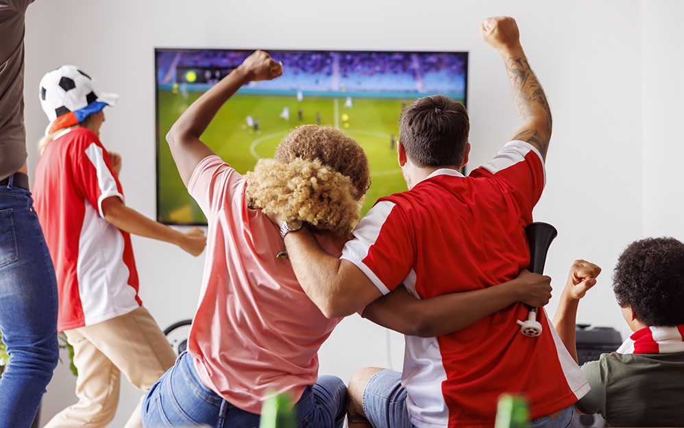 Group of young friends having fun watching football match on TV, drinking beer and cheering; football fans watching game at home celebrating after their team scoring a goal. Focus on the man in jersey